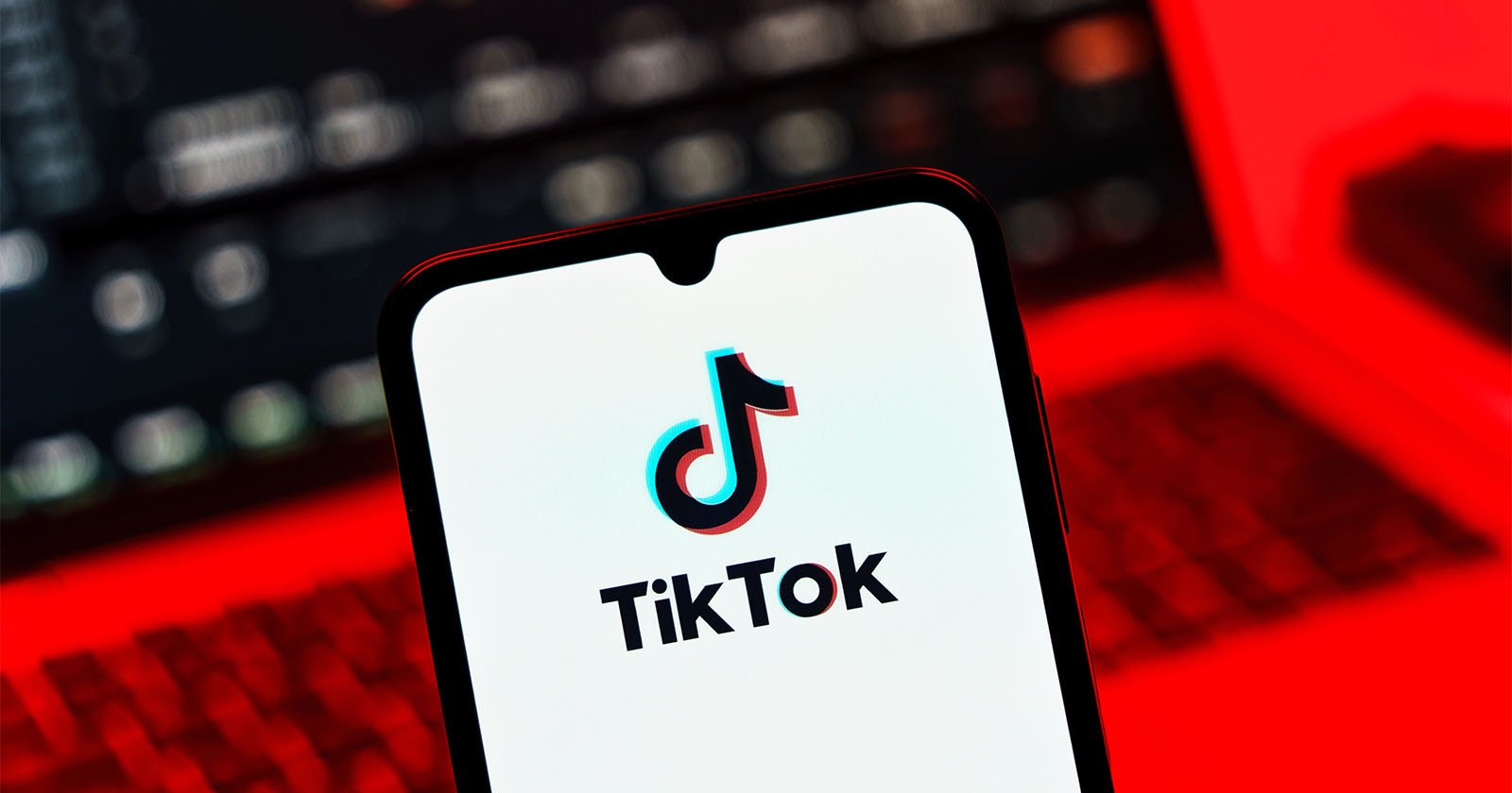 A smartphone displays the TikTok logo on its screen, with a blurred laptop in the background. The scene is illuminated by a red light. A smartphone displays the TikTok logo on its screen, with a blurred laptop in the background. The scene is illuminated by a red light.