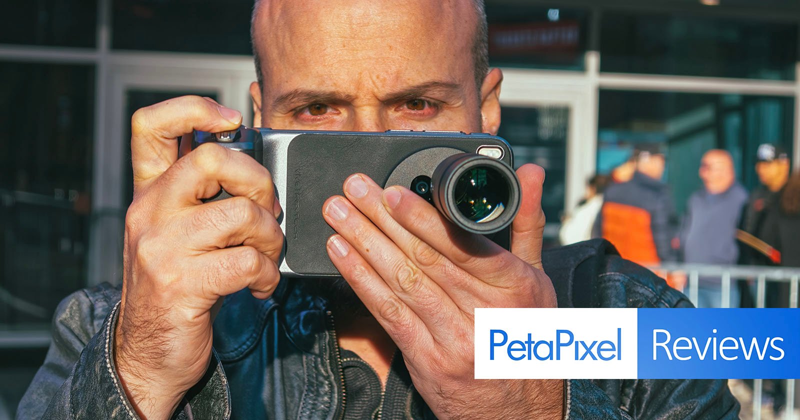 A man holds a smartphone with an attached camera lens, aiming it toward the viewer. The background is slightly blurred, showing people outdoors. The image features PetaPixel Reviews text in the bottom right corner. A man holds a smartphone with an attached camera lens, aiming it toward the viewer. The background is slightly blurred, showing people outdoors. The image features PetaPixel Reviews text in the bottom right corner.